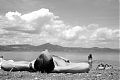 A photograph taken from ground level of a young man lying on a beach in Bracciano. A mountain and clouds can be seen in the distance, and another man lifting a baby out of the water can be seen in the middle-distance.